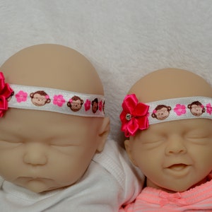 May include: Two baby headbands with a white background. Each headband features a white ribbon with monkey and pink flower patterns. A bright pink bow with a rhinestone center adorns each headband, adding a touch of elegance.