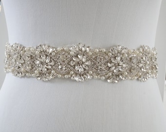 Op de afbeelding: Een witte parel bruidsgordel met een ontwerp van zilveren strass. De gordel is versierd met kleine parels en grote, heldere strass.