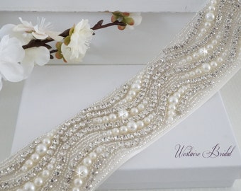 May include: A white satin bridal sash with a beaded design featuring pearls and rhinestones. The sash is on a white box with the text "Westaire Bridal".