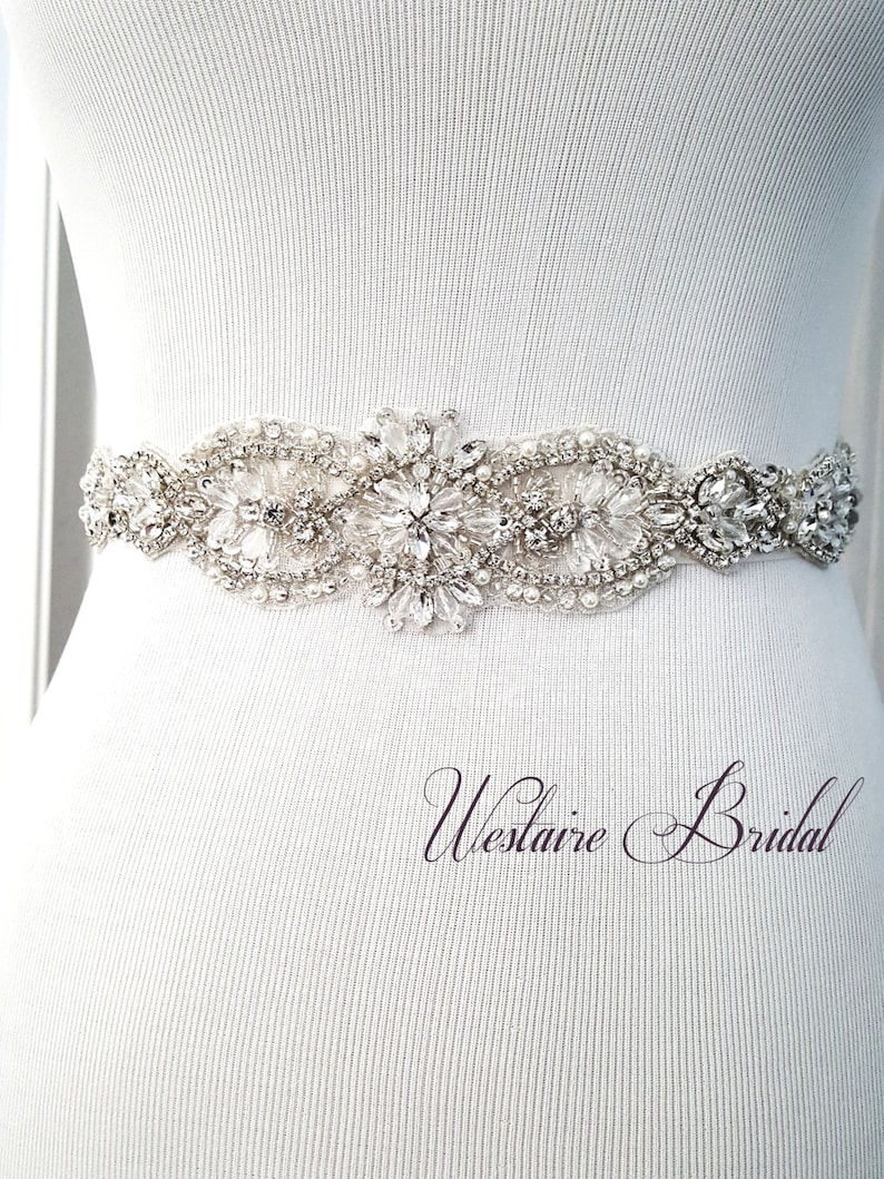 Op de afbeelding: Een witte bruidsgordel met kralen en strass met een bloemenpatroon. De gordel is op een wit mannequin.