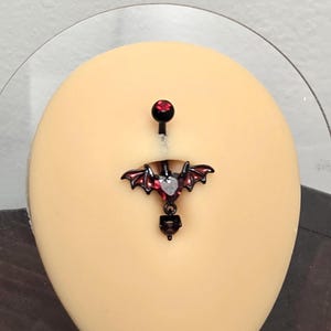 May include: A black belly button ring featuring a heart-shaped red gemstone centerpiece with attached black bat wings. The jewelry includes a small red gem on the top ball and a dangling black charm at the bottom, all set on a curved barbell post.