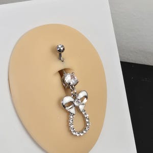 May include: A silver belly button ring with a clear gem on top and a dangling white bow design with a clear gem border. The top of the ring has a small, round gem.