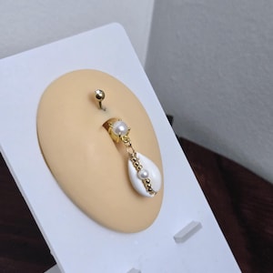 May include: A gold-colored belly button ring with a dangling white seashell pendant. The pendant is adorned with small pearls and gold beads. The ring is displayed on a tan-colored, oval-shaped form.