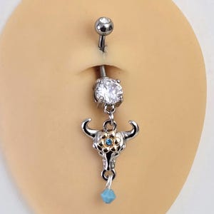 Pode incluir: Um piercing de umbigo prateado com uma gema transparente e um pingente de caveira de touro. A caveira de touro tem detalhes dourados e uma pequena flor azul. Um pequeno cristal azul pende da parte inferior do pingente.