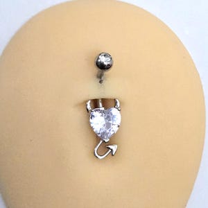 May include: A silver-toned belly button ring featuring a heart-shaped clear gemstone charm. The charm is designed with two small pointed horns at the top and a curved arrow-shaped tail at the bottom. A small clear gemstone is set in the top ball.