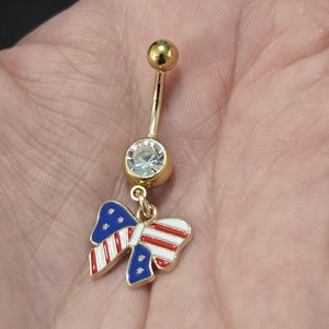 May include: Gold-toned belly button ring with a clear gem and a charm shaped like a bow. The bow charm features a patriotic design with red and white stripes and blue stars.