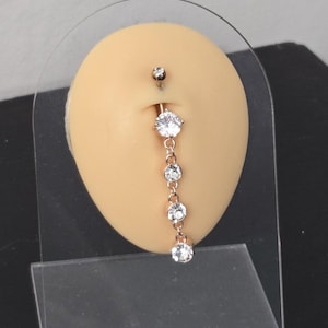 Puede incluir: Un piercing para el ombligo con un diseño colgante. La joya presenta un pequeño perno superior y una cadena de cuatro gemas redondas y brillantes. El metal tiene un tono dorado. La joya se exhibe sobre una forma de color beige.