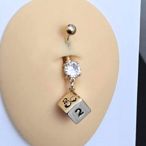 May include: A gold-colored belly button ring with a clear, round gem at the top and a larger, clear gem at the center. A small, gold-colored charm in the shape of a die is suspended from the center gem, with the number 2 visible.