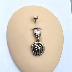 Könnte beinhalten: Ein Bauchnabelpiercing mit einem kleinen oberen Ball und einem Anhänger. Der Anhänger zeigt einen klaren Edelstein und ein bronzefarbenes Sonne- und Mond-Design. Der Schmuck wird auf einem hautfarbenen Hintergrund präsentiert.