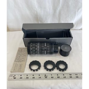 May include: A black cylindrical Stellarscope star finder with white printed months and time scales, accompanied by a matching storage box, three black circular rings, a small cap, and an instruction card, all positioned above a 12-inch ruler.