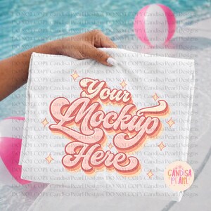 Op de afbeelding: Een witte rechthoekige handdoek met de tekst "Your Mockup Here" in een retro lettertype, in tinten roze en oranje. De handdoek wordt door een hand vastgehouden, met een vage achtergrond van een zwembad en strandballen.