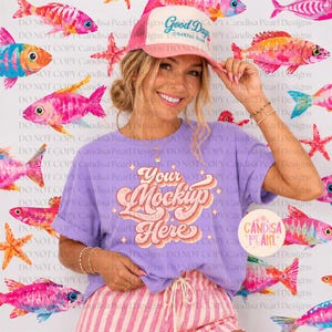 May include: A light purple t-shirt with a retro-style graphic that reads Your Mockup Here in pink and orange cursive text. The shirt is paired with pink and white striped shorts and a pink and white trucker hat with the text Good Days Coastal Club.