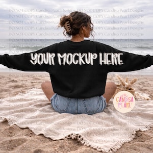 May include: Black long-sleeve sweatshirt with the text "YOUR MOCKUP HERE" in white. The sweatshirt is worn by a person sitting on a beach. A round tag with the text "Candisa Pearl" is visible.