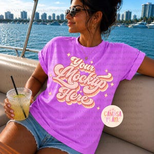 May include: A woman wearing a lilac t-shirt with the text "Your Mockup Here" in a retro design. She is wearing sunglasses and holding a drink with a straw. The background shows a cityscape and water.