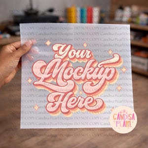 May include: A transparent sheet with the text "Your Mockup Here" in a retro font, in shades of pink and orange. The sheet is held by a hand, with a blurred background of various objects. The Candisa Pearl Designs logo is in the bottom right corner.