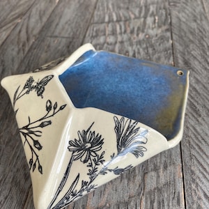 May include: A wall-mounted ceramic planter with a triangular envelope shape. The exterior is cream-colored with black line drawings of wildflowers and a butterfly. The interior features a deep blue glaze with a small hole for hanging on a wall.