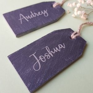 Può includere: Due etichette per segnaposto in ardesia con spago di iuta. Le etichette sono di colore grigio scuro con lettere bianche. I nomi "Audrey" e "Joshua" sono scritti sulle etichette.
