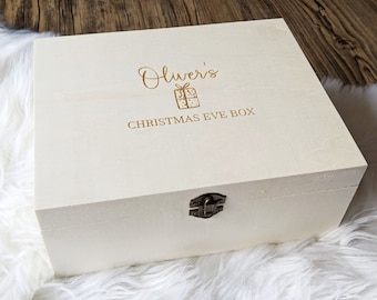 Puede incluir: Una caja rectangular de madera clara con tapa abatible y un cierre de metal color bronce. La parte superior de la caja presenta un texto en color dorado que dice Oliver's CHRISTMAS EVE BOX, junto a un pequeño gráfico de un regalo.