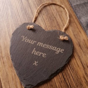 May include: A heart-shaped slate sign with the text "Your message here" and an "x" etched into the surface. A jute rope is attached for hanging. The sign is dark gray and rests on a wooden surface.