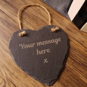 May include: A heart-shaped slate sign with the text "Your message here" engraved in the center. The sign has a natural rope hanger and a rustic aesthetic. The background is a wooden surface.