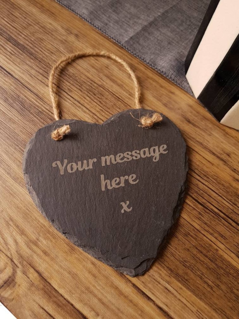 May include: A heart-shaped slate sign with the text "Your message here" engraved in the center. The sign has a natural rope hanger and a rustic aesthetic. The background is a wooden surface.