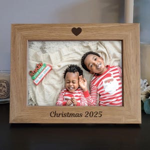 Könnte beinhalten: Hellbrauner Holzbilderrahmen mit einem Herz oben und dem Text "Christmas 2025" unten. Der Rahmen enthält ein Foto von zwei Kindern in rot-weiß gestreiften Pyjamas. Ein Schild mit der Aufschrift "Santa Stop Here We have cookies" ist ebenfalls zu sehen.