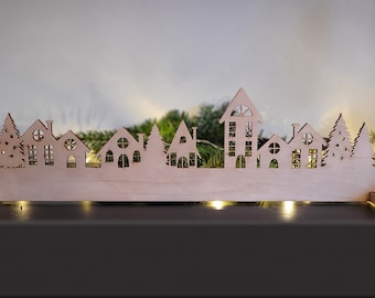 Puede incluir: Una silueta de pueblo navideño de madera con ventanas y árboles recortados. El pueblo está iluminado con luces blancas cálidas y se encuentra sobre una superficie negra.