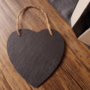 May include: A heart-shaped chalkboard with a natural rope hanger. The chalkboard is dark gray with a textured surface. The rope is a light brown color. The chalkboard is on a wooden surface.