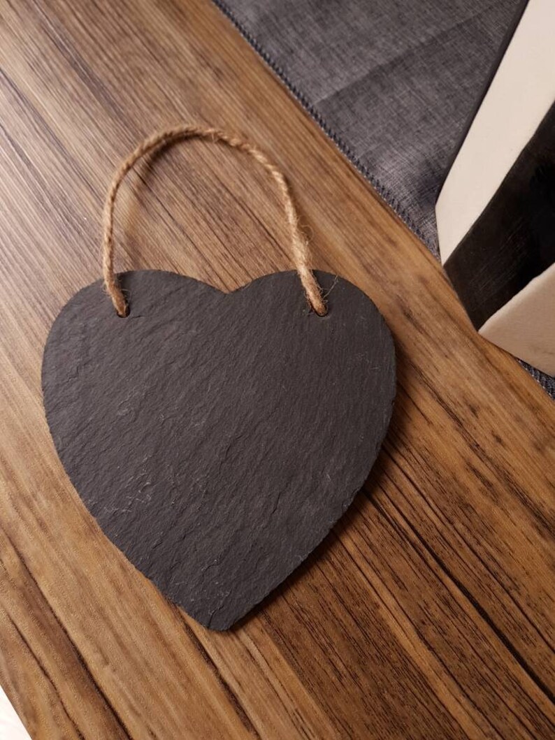 May include: A heart-shaped chalkboard with a natural rope hanger. The chalkboard is dark gray with a textured surface. The rope is a light brown color. The chalkboard is on a wooden surface.
