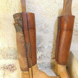 May include: Two antique wooden boot forms with leather coverings. The boot forms are made of light brown wood and have a natural finish. The leather coverings are brown and have a worn look. The boot forms are standing on a light brown surface.