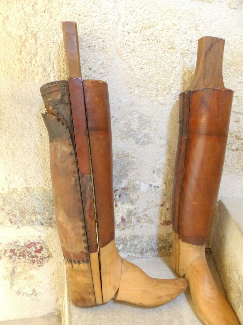 May include: Two antique wooden boot forms with leather coverings. The boot forms are made of light brown wood and have a natural finish. The leather coverings are brown and have a worn look. The boot forms are standing on a light brown surface.