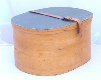 Può includere: Scatola ovale in legno con coperchio blu scuro e corpo marrone chiaro. Un cinturino in pelle marrone con fibbia assicura il coperchio. La scatola ha un aspetto rustico e antico.
