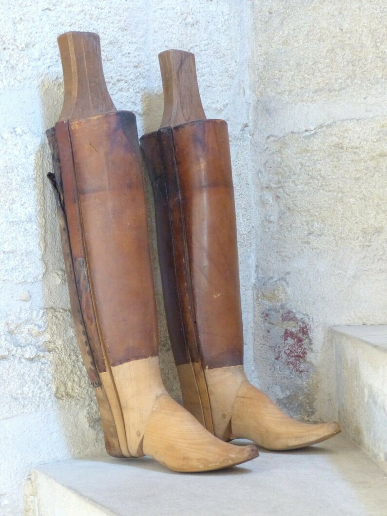 May include: Two wooden boot forms with brown leather coverings. The forms are made of wood and have a rounded toe. The forms are on a stone step.