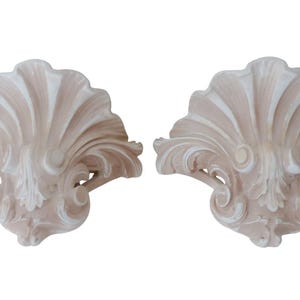 May include: A pair of decorative wall brackets in a light beige colour. The brackets are shaped like seashells with intricate floral details.