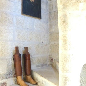 May include: Two brown leather boots with wooden shafts stand on a set of white stone stairs. The boots have a light brown leather toe and heel. A framed portrait of a man in a suit hangs on the wall behind the boots.