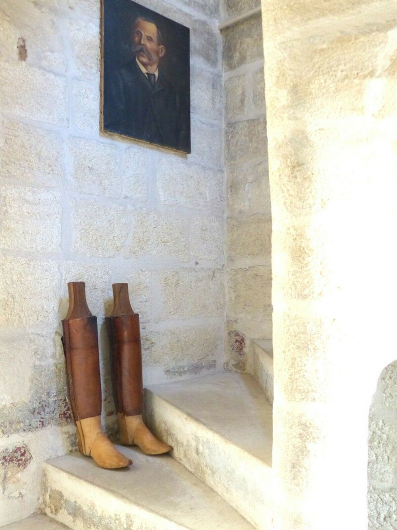 May include: Two brown leather boots with wooden shafts stand on a set of white stone stairs. The boots have a light brown leather toe and heel. A framed portrait of a man in a suit hangs on the wall behind the boots.