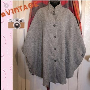 May include: A vintage gray and white houndstooth cape with a high collar and button closure. The cape has a classic design and is made of a woven fabric. The cape is a stylish outerwear option.