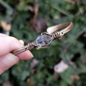 May include: A handcrafted bronze cuff bracelet featuring a faceted, gray crystal centerpiece. The crystal is secured with intricate wire wrapping. The bracelet has a hammered texture and is displayed against a blurred green background.