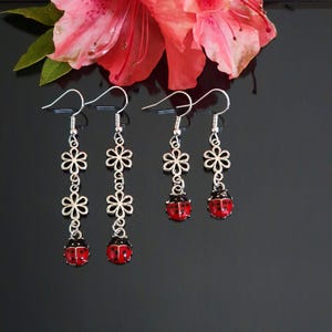 May include: Silver-coloured drop earrings with a floral design and red ladybug charms. The earrings have a hook closure and are displayed against a black background with a pink flower.