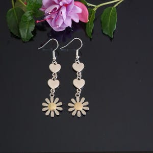May include: A pair of silver-coloured drop earrings. Each earring has three heart-shaped charms and a daisy pendant with a gold-coloured centre. The earrings are set against a black background, with a pink and purple flower.