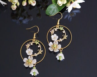 May include: Gold hoop earrings with white and green flower accents. The flowers are made of a pearlescent material and have a delicate, feminine design.