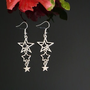 May include: A pair of silver star earrings. Each earring has three stars of different sizes, suspended from a hook. The earrings are set against a dark background, with a red flower in the top left corner. The earrings are a stylish accessory.