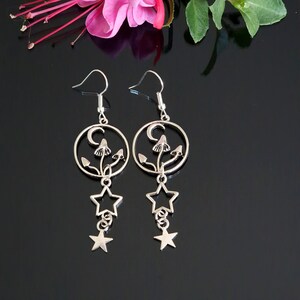 May include: Silver-coloured drop earrings with a celestial design. Each earring features a circular frame with a crescent moon and mushroom motif, with a dangling star charm. The earrings have a hook closure.