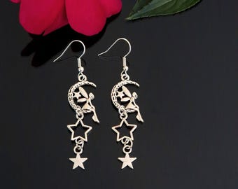 May include: Silver-toned dangle earrings featuring a crescent moon design with a seated figure and dangling stars. The earrings have a hook closure and are displayed against a black background.