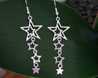 May include: A pair of silver-colored dangle earrings. Each earring features a large star at the top, with three smaller stars dangling below. The earrings are suspended from a hook. The background is a dark green leaf.