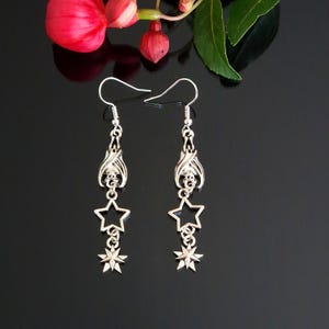 May include: A pair of silver-coloured drop earrings. Each earring has a bat design at the top, followed by a star and a smaller, multi-pointed star charm. The earrings hang from a simple hook. The background is black, with a red flower and green leaves.