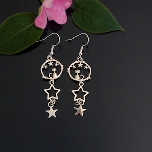 May include: A pair of silver-toned drop earrings featuring a circular charm with a cat silhouette and small stars, dangling below are two additional open star shapes. The earrings hang from standard silver-toned metal fishhook ear wires.