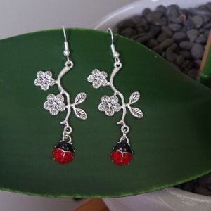 May include: Silver dangle earrings with a floral design and a ladybug charm. The earrings are on a green leaf background.