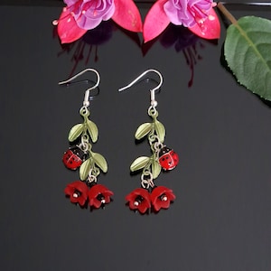 May include: Dangle earrings with a ladybug theme. The earrings feature red ladybugs, red flower buds, and green leaves. They are suspended from silver-coloured hooks. The background is black.