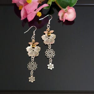 May include: A pair of silver-toned drop earrings featuring a small bee charm with yellow and black stripes resting on a five-petaled flower. Below the flower hangs a circular geometric filigree charm and a tiny five-petaled flower charm.